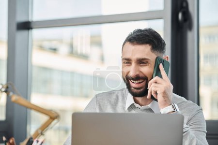 Ein Mann sitzt vor einem Laptop und telefoniert mit einem Handy, vertieft in seine Arbeit in einem geschäftigen Büro.