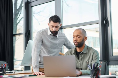 Two men in a corporate office are focused on a laptop screen, actively engaged in a discussion or project analysis, diversity and inclusion