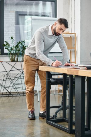Un hombre centrado en su portátil, de pie en un escritorio en una oficina ocupada, trabajando en un proyecto integral de la cultura corporativa.