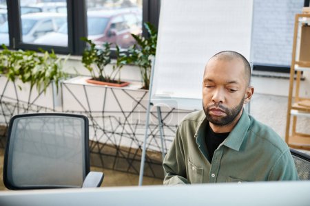 Ein behinderter afrikanisch-amerikanischer Mann mit Myasthenia gravis sitzt an einem Schreibtisch vor einem Computerbildschirm in einem geschäftigen Büroumfeld