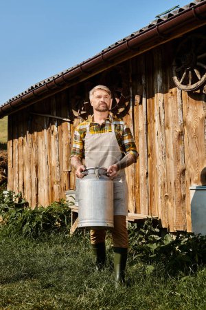 appealing farmer in casual attire with tattoos holding milk churn and looking away next to house