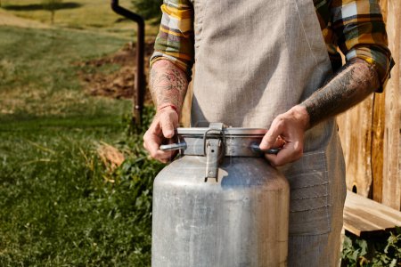 vista recortada de agricultor adulto con tatuajes en brazos sosteniendo batido de leche de metal mientras está en la aldea