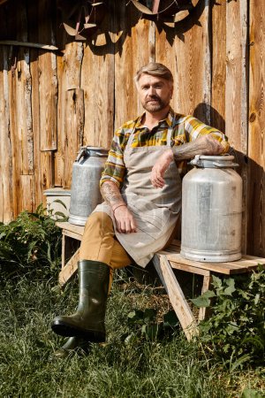 good looking farmer in casual attire with tattoos sitting with milk churns and looking at camera