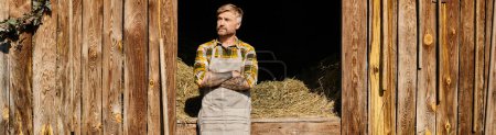 Téléchargez les photos : Beau fermier en tenue décontractée à l'aide d'une fourche tout en travaillant avec du foin dans le village, bannière - en image libre de droit