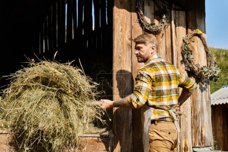 Foto de Atractivo agricultor dedicado en atuendo casual usando horquilla mientras trabaja con heno en la aldea - Imagen libre de derechos