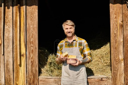 joyous handsome farmer with tattoos holding homemade cheese in his hands and smiling at camera