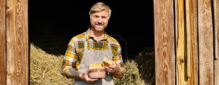 Foto de Alegre granjero guapo con tatuajes sosteniendo queso casero en las manos y sonriendo a la cámara, pancarta - Imagen libre de derechos