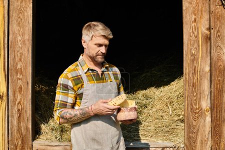 hombre guapo con tatuajes en brazos en atuendo casual sosteniendo queso y mirando hacia otro lado mientras está en la granja