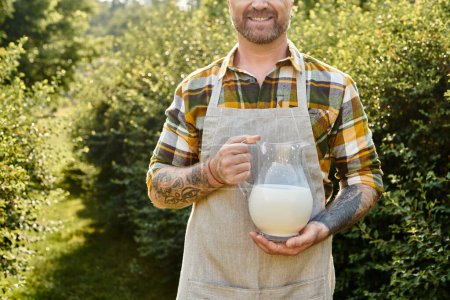 Foto de Vista recortada de alegre granjero con tatuajes sosteniendo frasco de leche fresca y sonriendo felizmente - Imagen libre de derechos