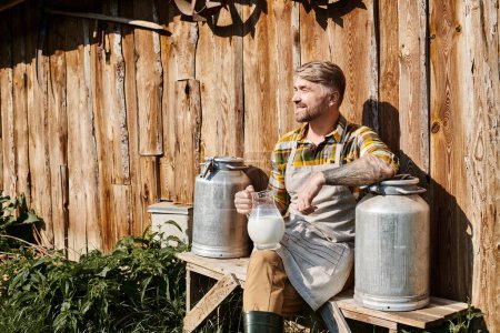 Téléchargez les photos : Joyeux fermier attrayant assis près de sa maison avec pot et barattes de lait et souriant loin - en image libre de droit