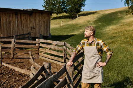 bel homme en tenue décontractée avec des tatouages posant à côté de la clôture et du fumier à la ferme et regardant loin