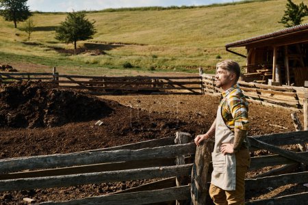 handsome man in casual attire with tattoos posing next to fence and manure on farm and looking away