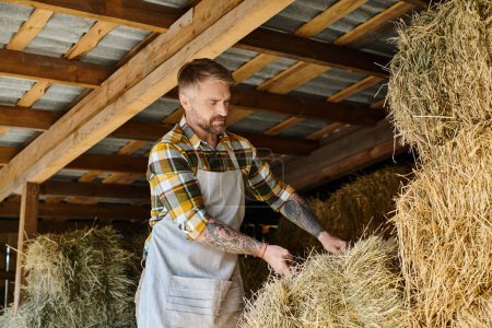 Foto de Atractivo hombre dedicado con barba y tatuajes trabajando con fardos de heno mientras está en su granja - Imagen libre de derechos