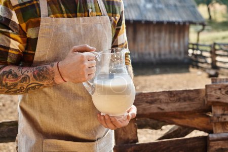 abgeschnittene Ansicht eines hart arbeitenden Mannes mit Tätowierungen, der auf seinem Bauernhof ein großes Glas mit frischer Milch hält