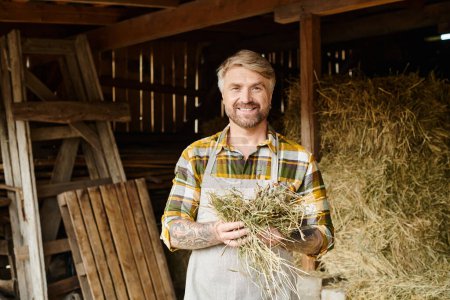 Foto de Alegre granjero bien parecido con barba y tatuajes sosteniendo un poco de heno y sonriendo a la cámara en la granja - Imagen libre de derechos