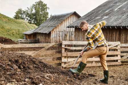 Photo for Hard working attractive farmer with beard and tattoos using pitchfork while working with manure - Royalty Free Image