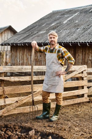 handsome farmer with tattoos using pitchfork while working with manure and looking at camera