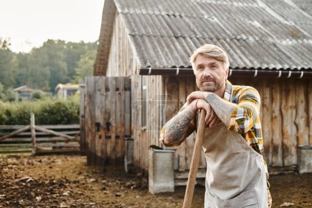 trabajador atractivo granjero con barba y tatuajes usando horquilla mientras se trabaja con estiércol