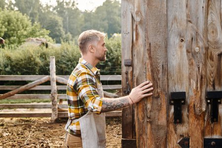 atractivo hombre dedicado con barba y tatuajes en sus brazos cerrando puertas de granero mientras está en la granja