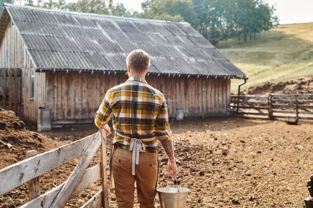 Rückansicht eines erwachsenen fleißigen Mannes mit Tätowierungen auf den Armen, der Eimer mit Milch hält, während er auf dem Bauernhof ist