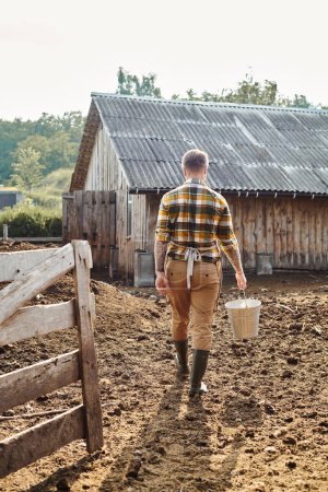back view of adult hardworking man with tattoos on arms holding bucket with milk while on farm