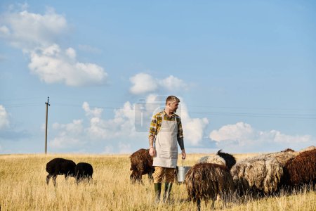 attraktiver, hart arbeitender Bauer mit Bart, der Eimer mit Milch hält, umgeben von Schafen und Lämmern