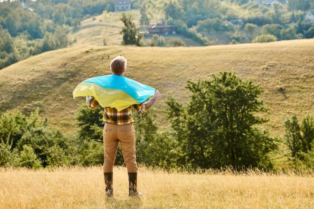 back view of adult Ukrainian farmer with tattoos on arms posing with national flag, scenic landscape