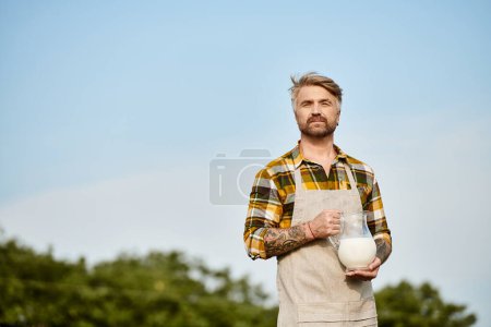 granjero moderno de buen aspecto con barba y tatuajes sosteniendo frasco de leche fresca y mirando a la cámara