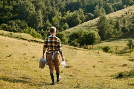 Photo for Back view of hard working modern farmer with tattoos walking with bucket and jar of milk in hands - Royalty Free Image
