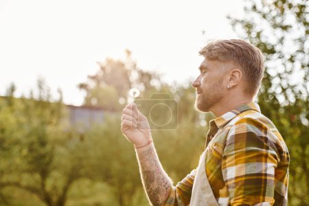 alegre atractivo hombre con tatuajes soplando en diente de león mientras se enfría en la granja a la luz del sol