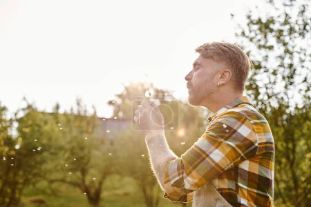 fröhlich attraktiver Mann mit Tätowierungen, die auf Löwenzahn wehen, während er auf einem Bauernhof im Sonnenlicht chillt