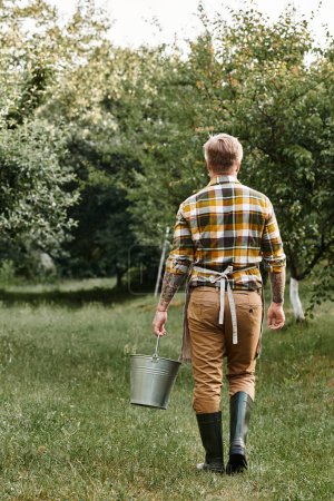 vertikale Aufnahme eines hart arbeitenden Mannes mit Tätowierungen, der im Garten arbeitet und einen Metalleimer in der Hand hält
