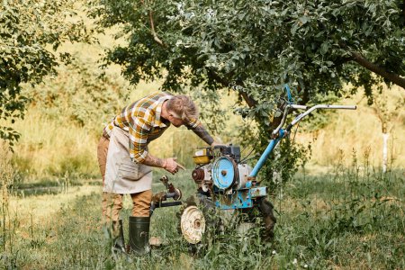 schöner bärtiger Mann in Freizeitkleidung mit Tätowierungen mit Rasenmäher im Garten auf seinem Bauernhof