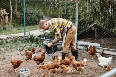 good looking joyous man with tattoos feeding chickens in their aviary while on his farm in village