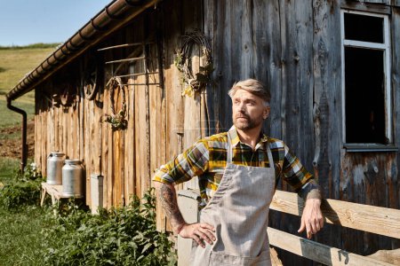 Foto de Atractivo barbudo moderno agricultor en traje casual con tatuajes posando y mirando hacia otro lado en la aldea - Imagen libre de derechos