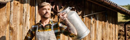 Photo for Appealing man in casual attire with tattoos posing with milk churns and looking away, farmer, banner - Royalty Free Image