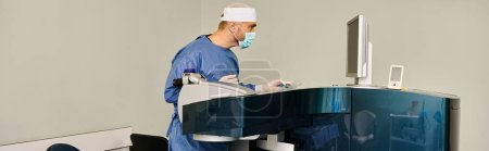 A man in a surgical suit stands before a desk with a monitor.