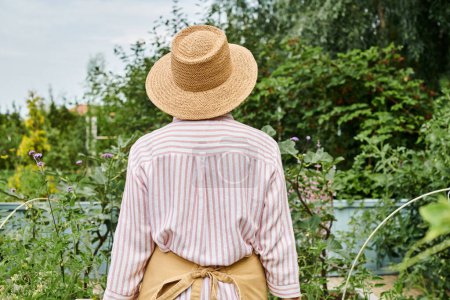 Rückseite der reifen Frau mit Strohhut posiert in ihrem lebhaften Garten, während sie dort arbeitet