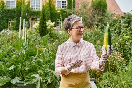 attractive merry mature woman in casual attire with glasses holding fresh zucchini in her garden