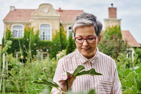 atractiva mujer madura feliz con gafas trabajando en su jardín verde vívido y sonriendo alegremente