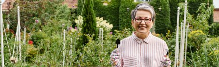 Photo for Jolly mature woman with glasses holding gardening tools in hands and smiling at camera, banner - Royalty Free Image