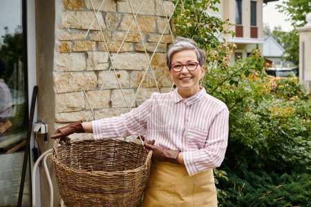 cheerful mature woman holding big straw basket and smiling at camera near her house in England