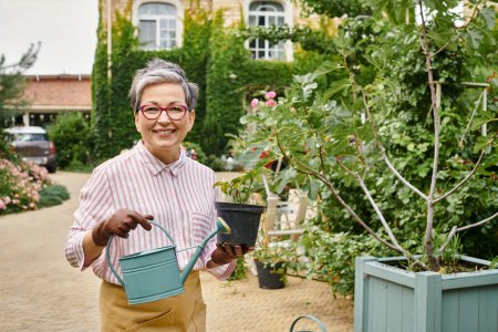 ansprechende freudige reife Frau mit Pflanze in der Hand, Gießkanne und Topf in die Kamera lächelnd, England