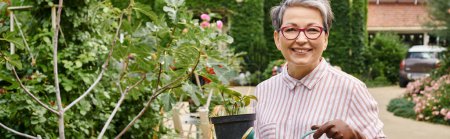 joyful mature woman taking care of plant in pot in garden in England and smiling at camera, banner