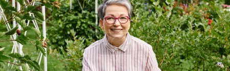 joyful beautiful mature woman in casual attire with glasses smiling at camera in her garden, banner