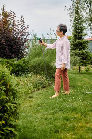 Téléchargez les photos : Belle femme mature gaie avec des lunettes en utilisant un tuyau pour arroser ses plantes vivantes dans son jardin - en image libre de droit