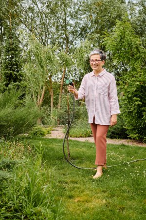 gut aussehende, reife Frau mit Brille, die ihre lebendigen Pflanzen in ihrem Garten mit einem Schlauch wässert