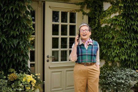 mature cheerful elegant woman with glasses and short hair talking by her phone near house in England