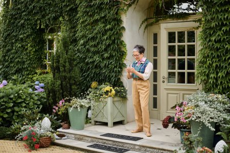 kultivierte, reife Frau mit Brille, die in der Nähe ihres Hauses in England auf ihr Handy schaut
