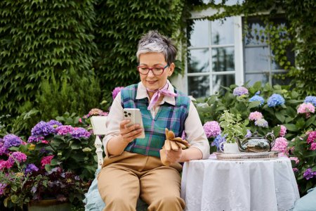 reife, fröhliche Frau, die zur Teezeit im Garten sitzt und in der Nähe ihres Hauses in England auf das Telefon schaut